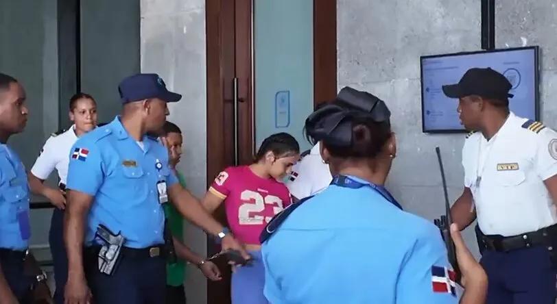 Security officers in blue uniforms escort a woman in a pink shirt toward a doorway near a building entrance.