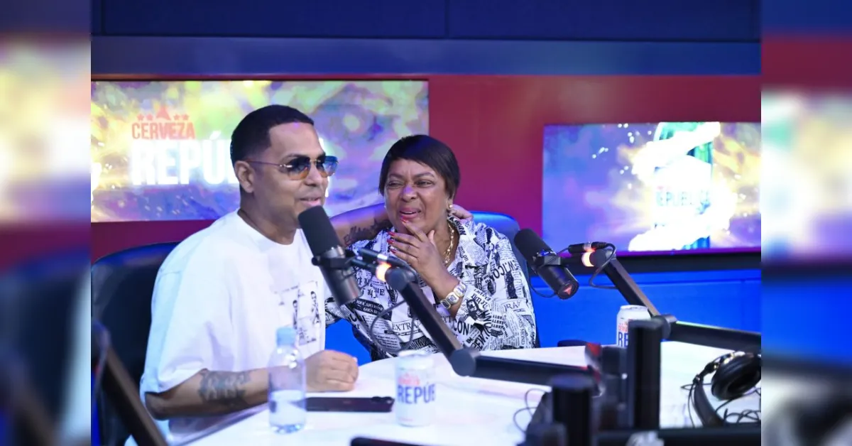 Two radio hosts chat at a studio desk, speaking into microphones as they smile at each other.