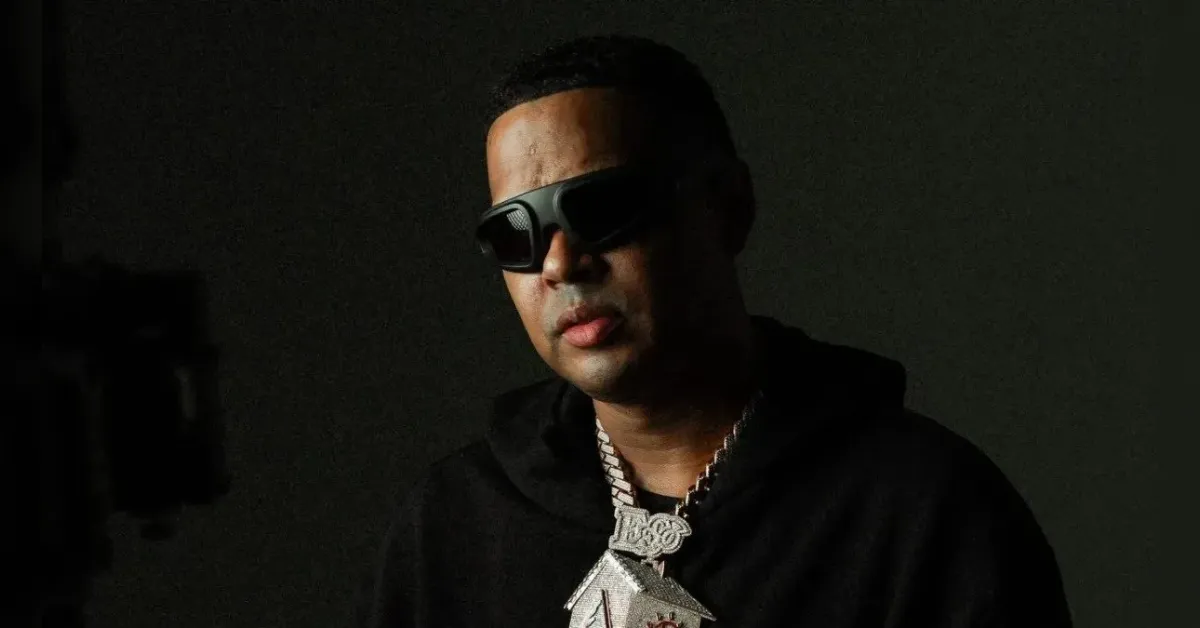 Portrait of a man in a black hoodie and dark sunglasses, wearing a thick silver chain with a large pendant in a moody, low-light setting.