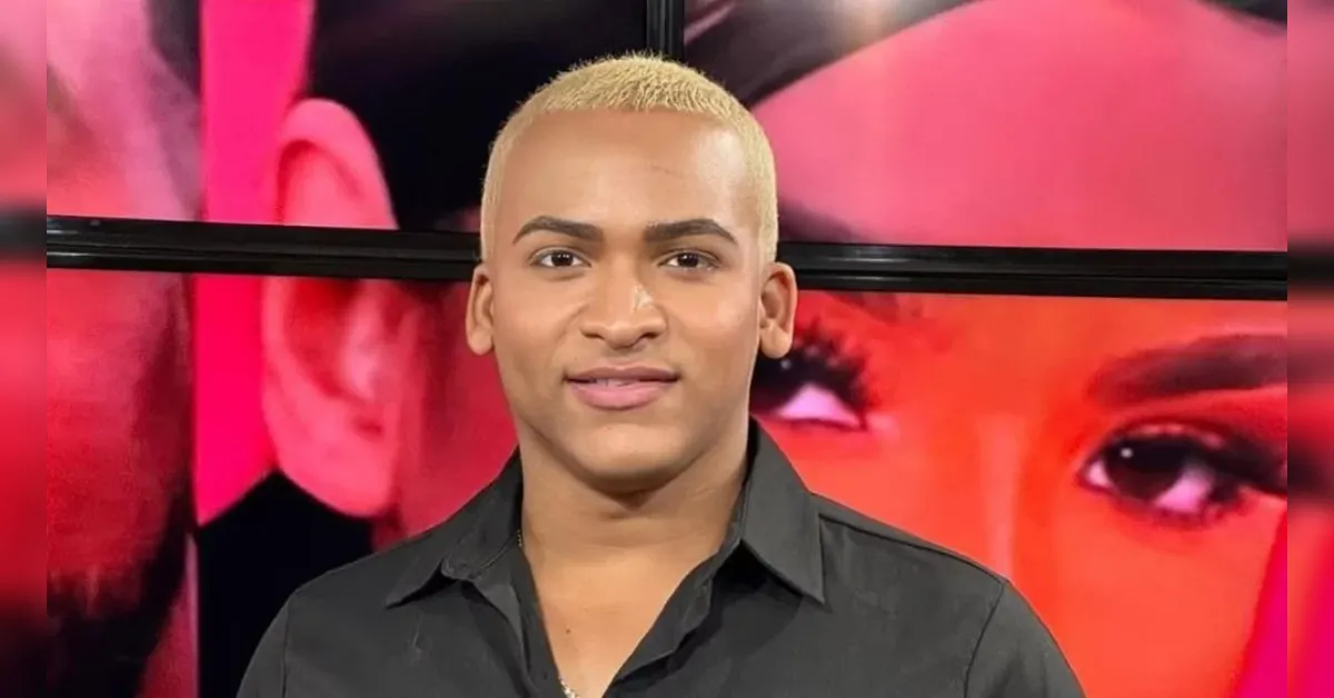 Portrait of a person with short platinum blonde hair wearing a black shirt, standing in front of a vibrant studio backdrop with pink and red tones.