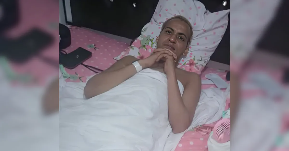 Person lying in bed under a white blanket, hands clasped under the chin, looking at the camera.