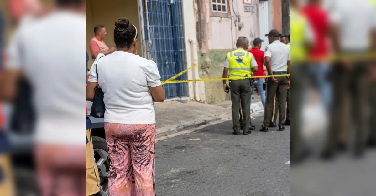 Street scene with yellow caution tape; people stand and talk near a doorway, including officers in reflective vests. Woman in white top and pink patterned pants seen from behind in foreground.