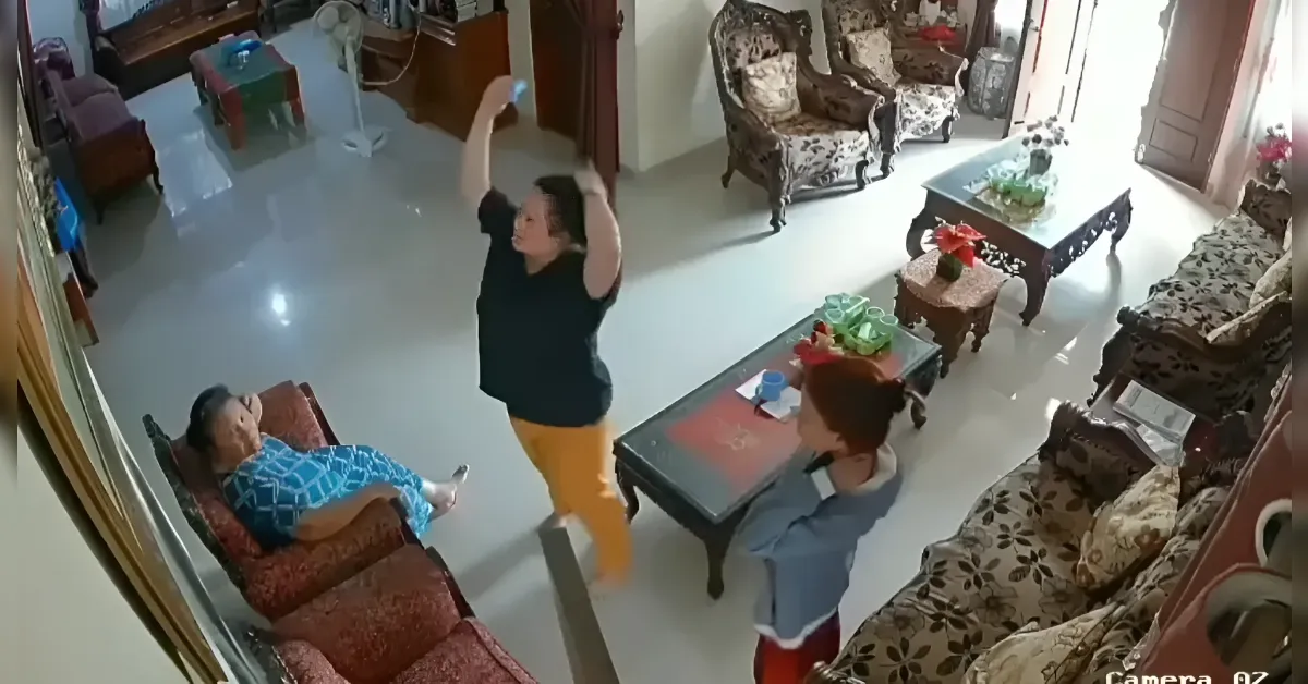 Living room scene with two older people seated on patterned chairs; a younger woman in black top and orange pants raises her arms as another person in blue stands near a coffee table.
