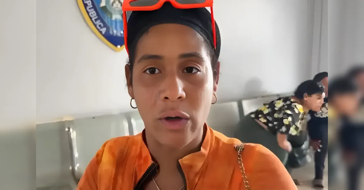 Close-up of a woman in an orange jacket speaking to the camera, with red sunglasses on her head and hoop earrings visible.