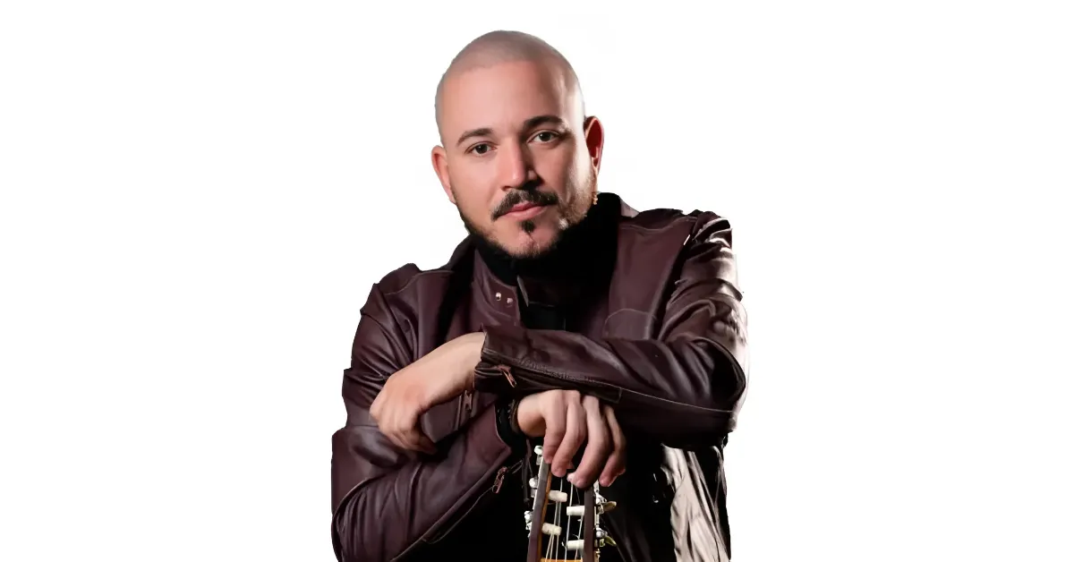 Man with a shaved head and beard leaning on a guitar, wearing a brown leather jacket, looking at the camera.
