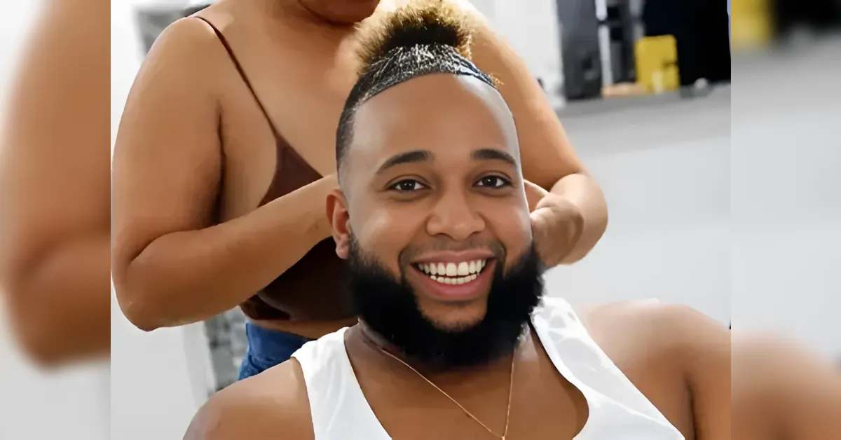 Smiling man with a fresh mohawk and full beard being styled by a barber in a shop, mid-session.