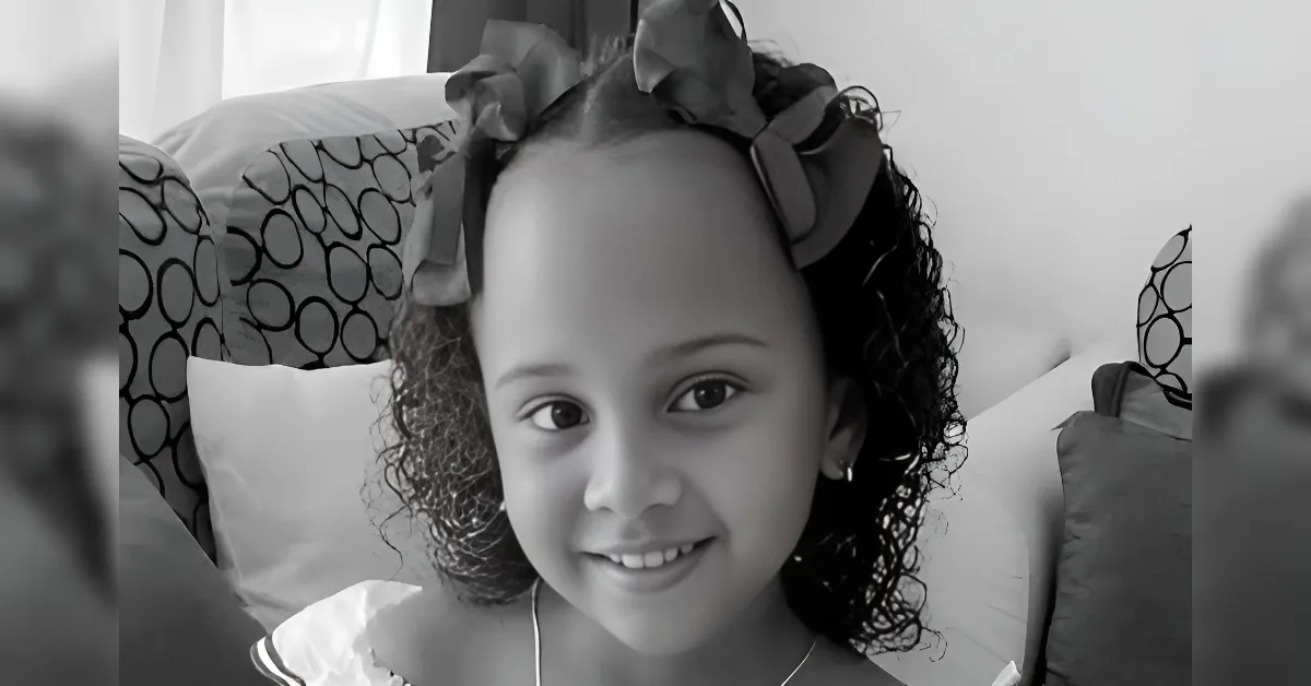 Smiling young girl with a bow headband, sitting on a couch with patterned cushions indoors.