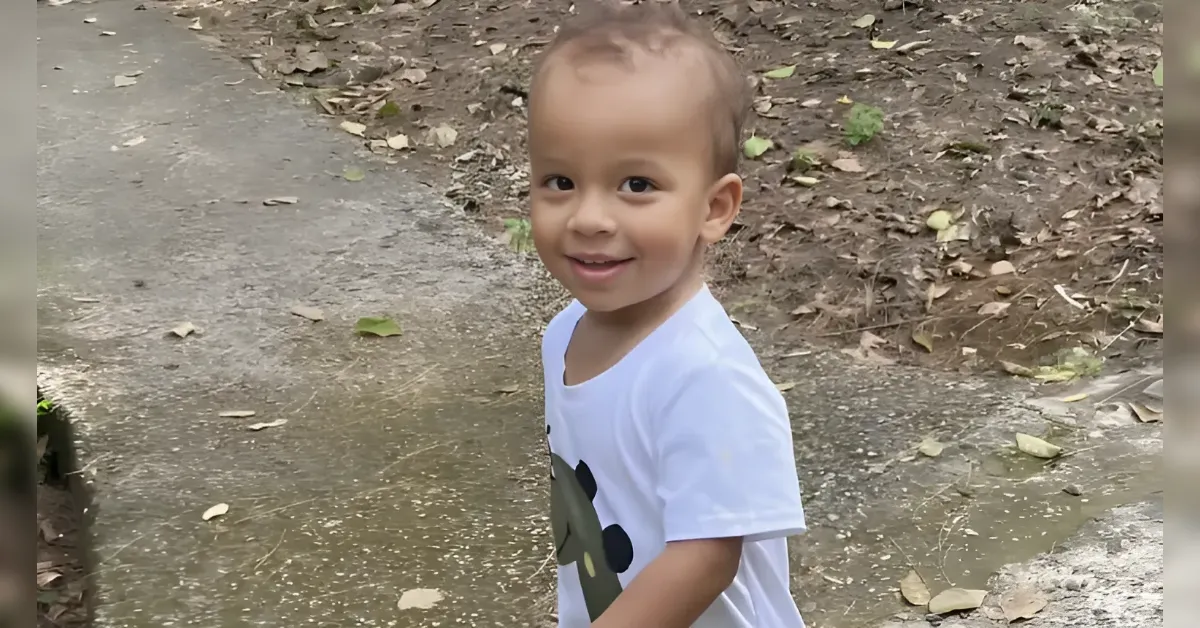 Smiling toddler in a white shirt standing on a leaf-strewn path outdoors.