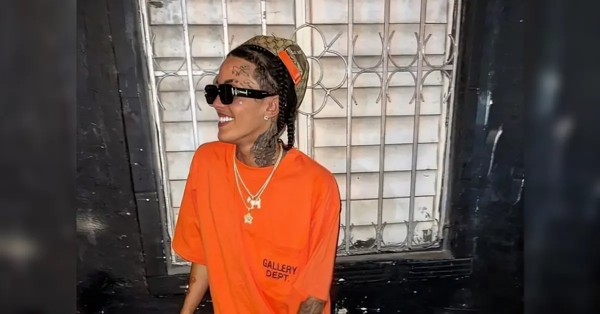 Smiling person in an orange T‑shirt and black sunglasses stands near a barred window. Tattoo on neck visible under braids.