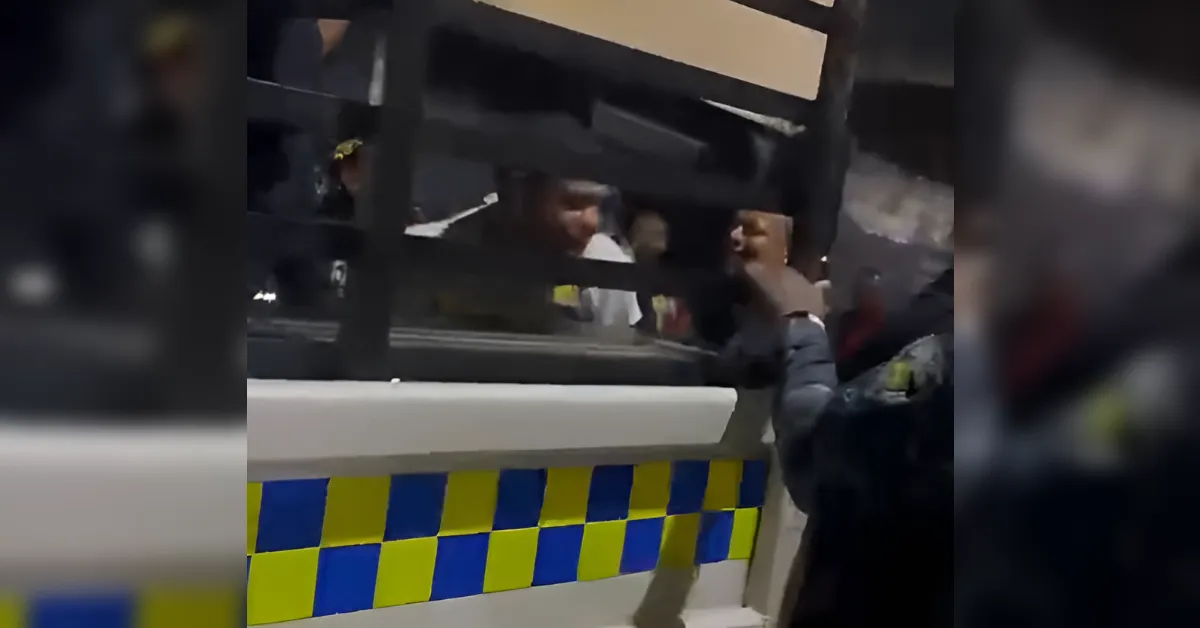 Nighttime scene of a police van with blue-and-yellow checkered markings and a person near a window.
