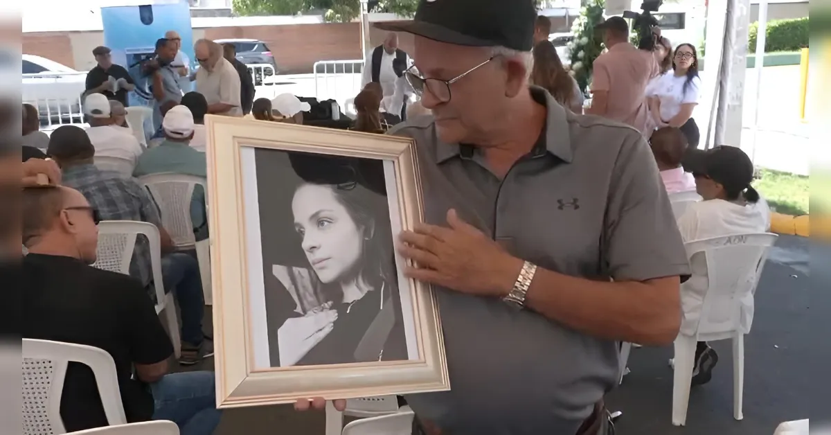 Man in a gray polo presents a framed black-and-white portrait of a woman at an outdoor memorial gathering.