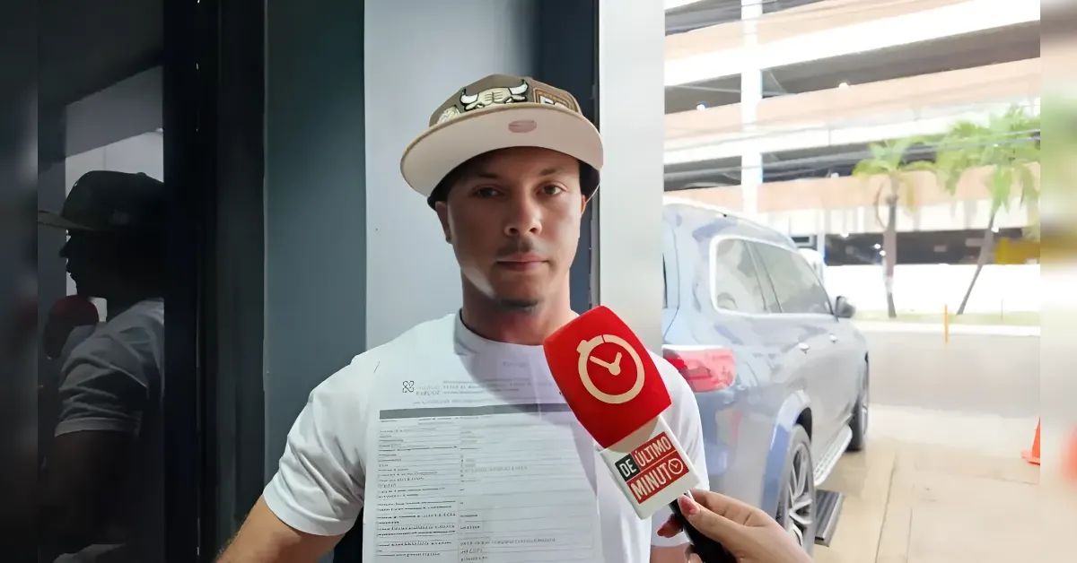 Man wearing a beige cap being interviewed by a reporter, holding a printed document beside a parked car.