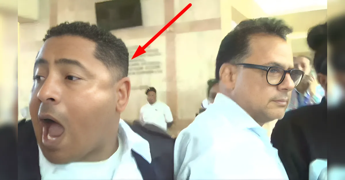 Two men in a crowded indoor space; the man on the left has his mouth open in surprise while the man on the right wears glasses and looks away, with a red arrow pointing to the left side of the image.