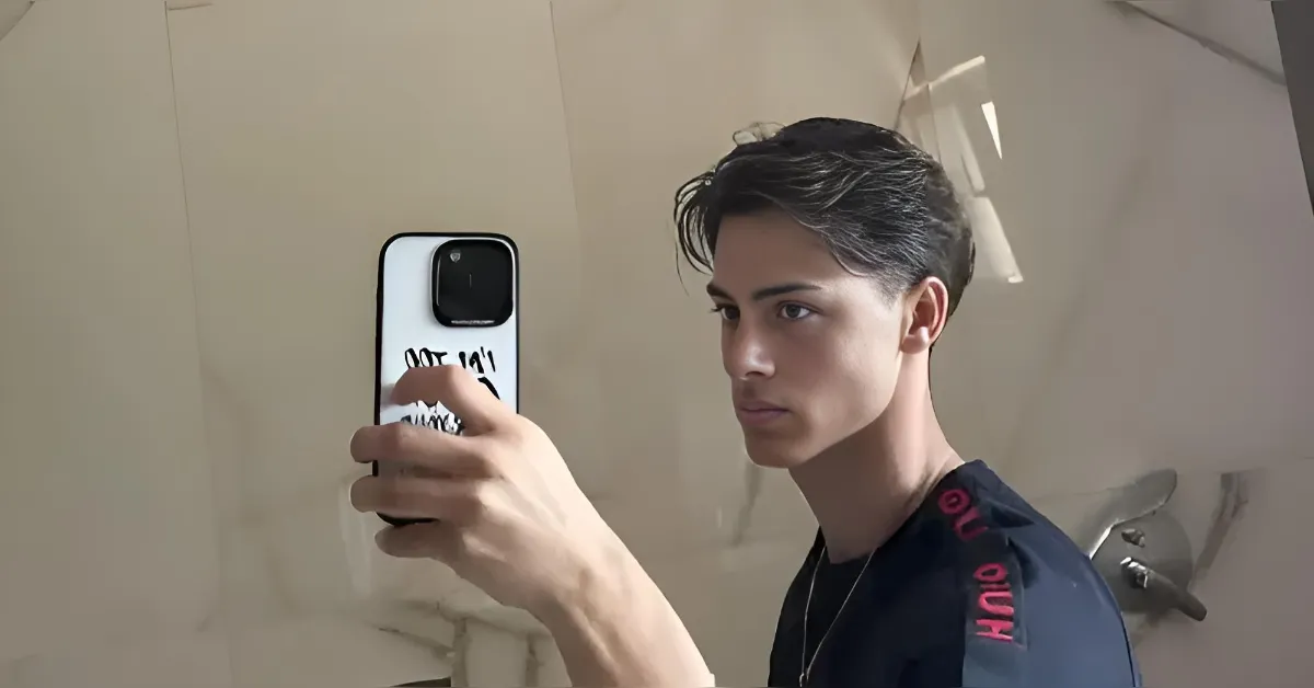 Young man taking a mirror selfie in a bathroom, holding a smartphone with a large rear camera bump.