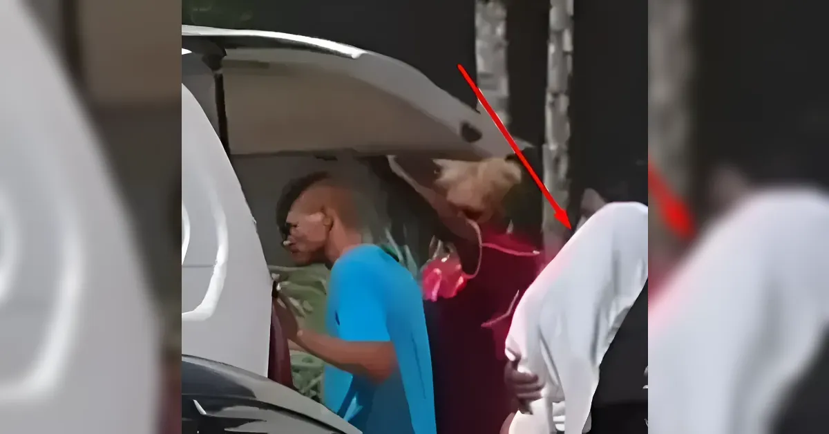 Several people stand around an open car trunk, with one person in a bright blue shirt leaning toward the vehicle while others are nearby in red and white clothing.