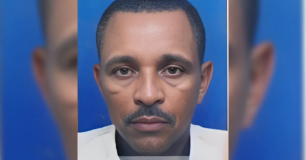 Close-up portrait of a man with short hair and a mustache, wearing a light-colored shirt, against a solid blue background with a neutral expression.