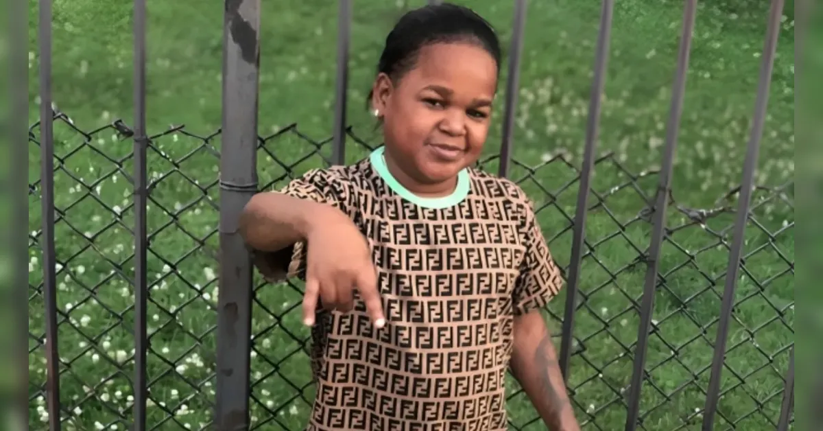 Child wearing a tan shirt with a repeating pattern, leaning on a metal fence and pointing at the camera.