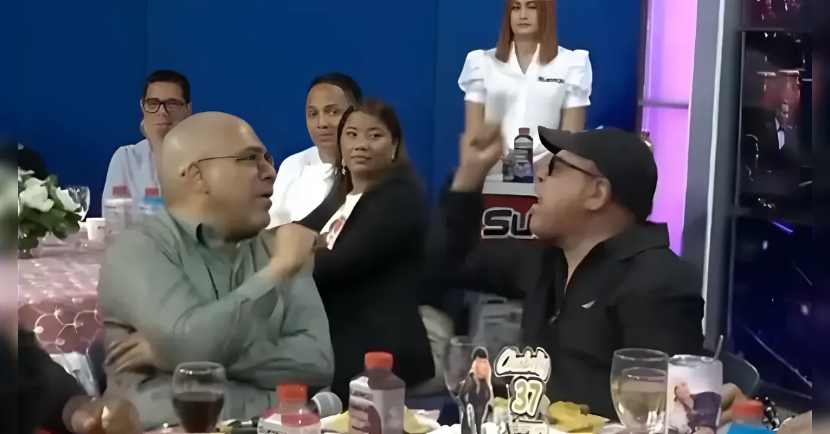 Two men argue across a dining table while others look on in a banquet setting.