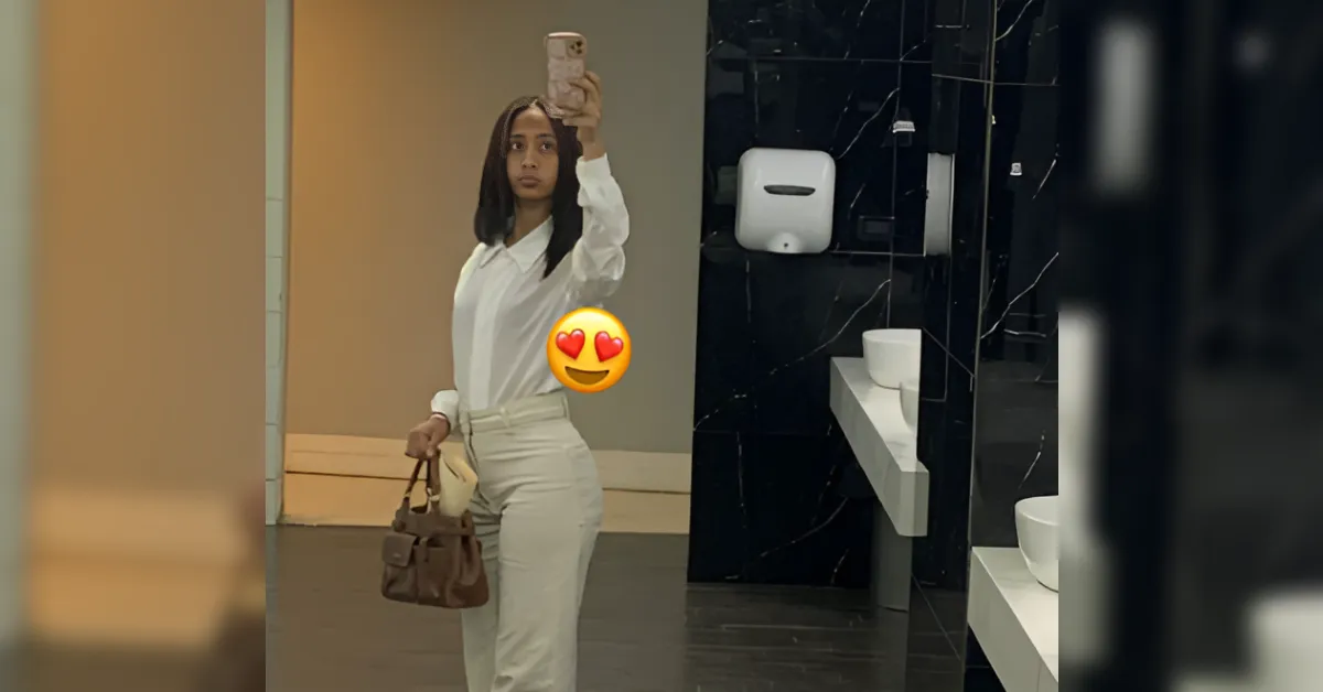 Woman in a white blouse and cream pants takes a mirror selfie in a bathroom, holding a brown handbag.