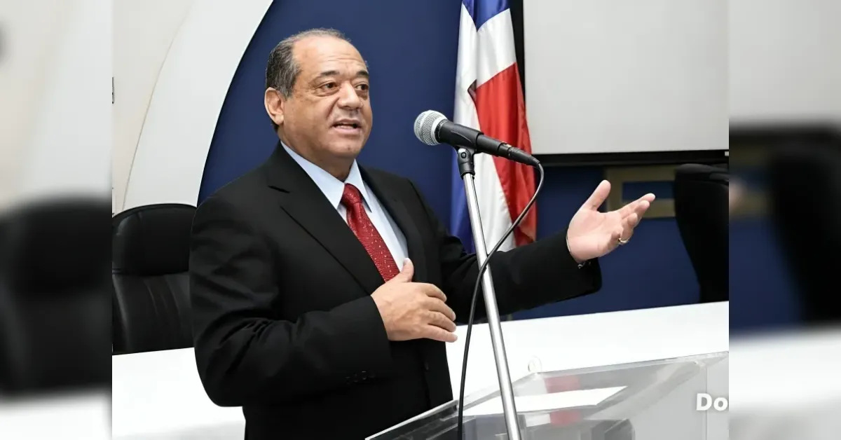 Man in a dark suit and red tie speaks at a clear lectern, gesturing with one hand in front of a microphone and a flag in the background.