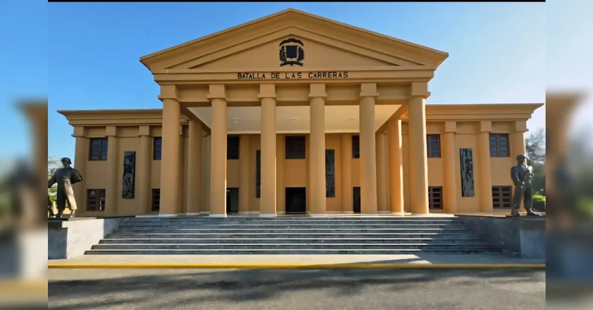 Front view of a tan neoclassical building with tall columns, wide steps, and statues on either side; inscription 'Batalla de Las Carreras' above the entrance.