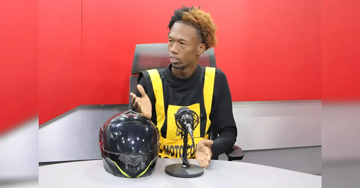 Man in a yellow safety vest speaks into a microphone at a podcast studio, with a motorcycle helmet on the table beside him against a red backdrop.