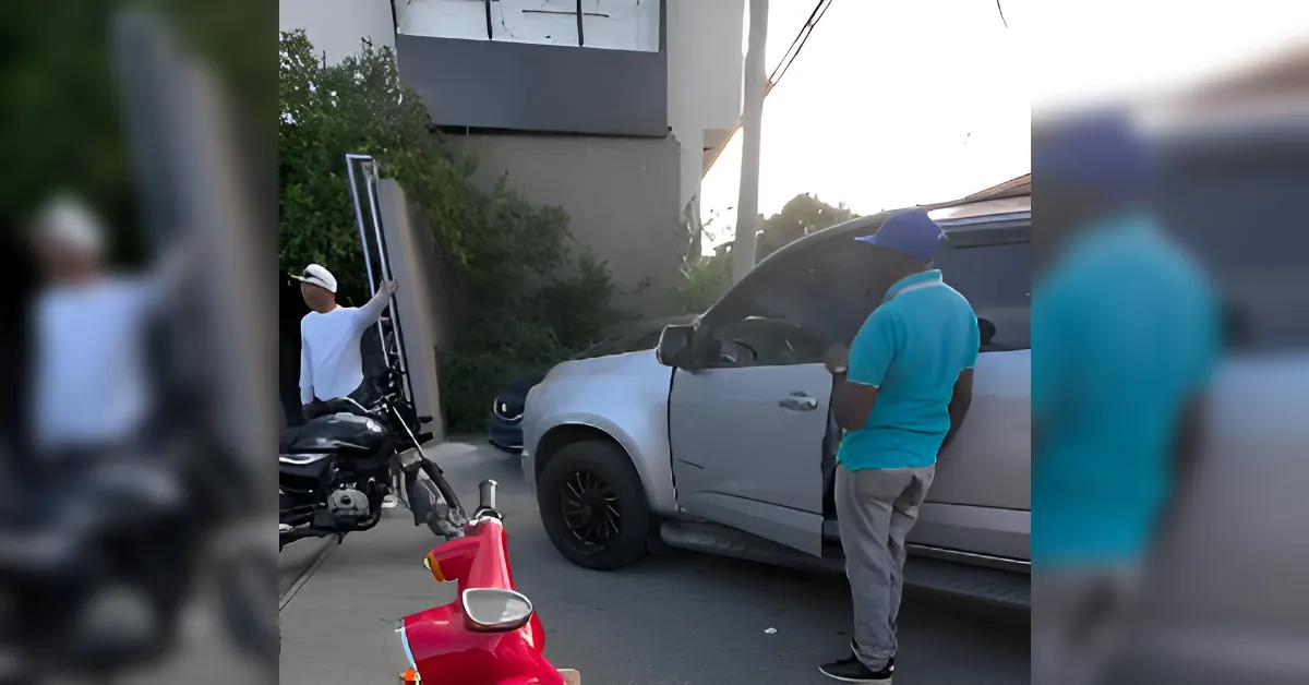 Two men stand by a gray pickup truck with its hood open while a motorcycle and a red scooter sit nearby on the street.