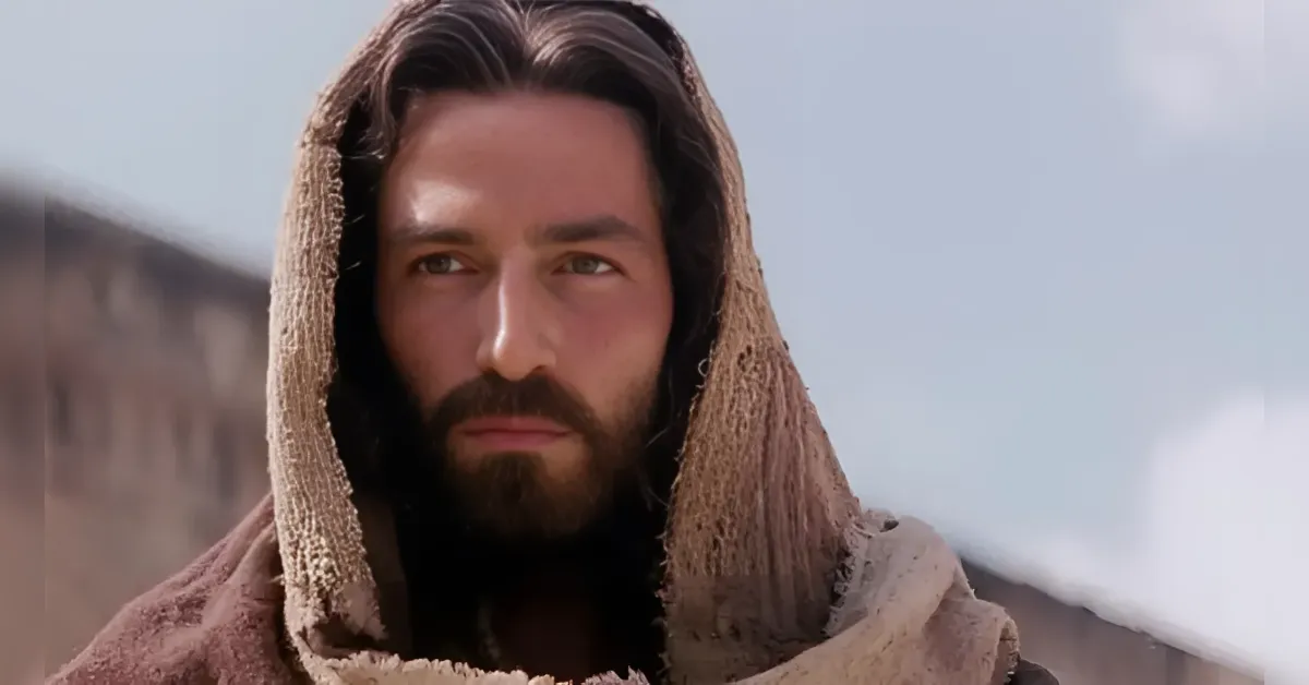 Close-up of a bearded man in a rough brown hooded cloak, gazing into the distance against a pale sky.