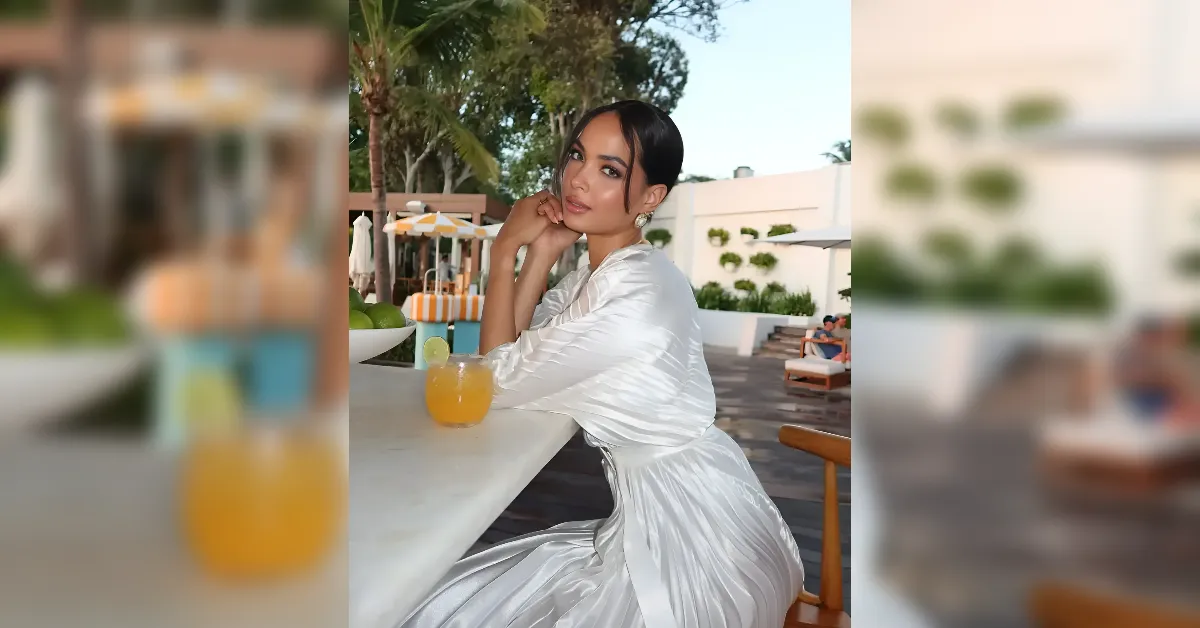 Woman in a white satin dress sits at a marble bar by a pool, resting her chin on her hands with an orange juice nearby.