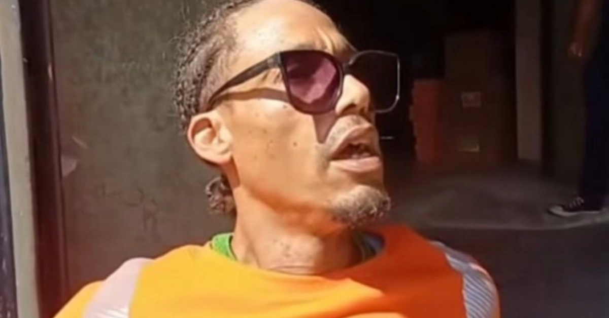 Man wearing an orange shirt and large sunglasses, looking upward to the left in an urban outdoor setting.