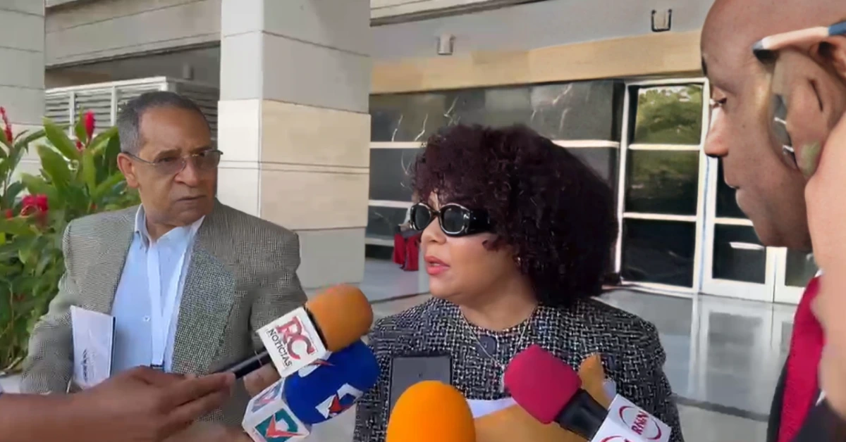 Woman in black sunglasses answering questions from reporters outside a modern building, microphones held close to her.