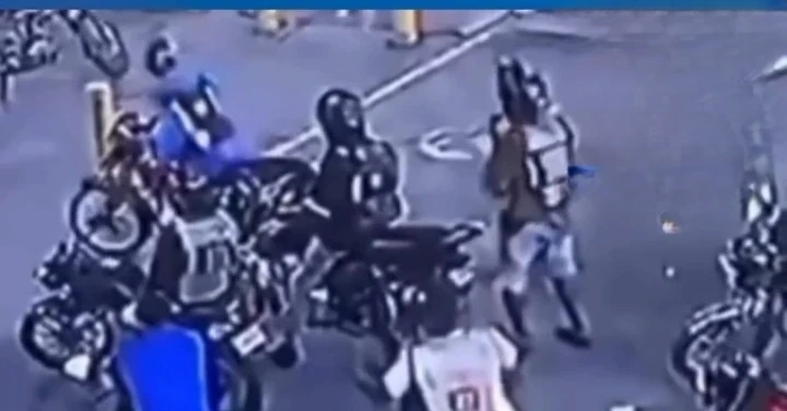 Group of people gathered around parked motorcycles on a street, some wearing helmets, seen from a surveillance camera angle.