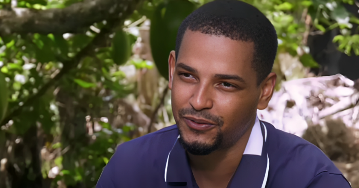 Man with short hair and goatee wearing a dark blue polo outdoors, looking to the side, with greenery in the background.