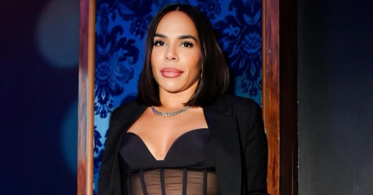 Portrait of a woman with shoulder-length dark hair, wearing a black blazer over a sheer bustier, and a silver necklace, standing in a doorway against a blue patterned wall.