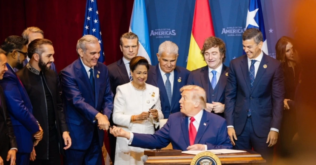 Donald Trump sits at a desk signing a document as a group of officials and world leaders look on. Flags stand in the background.