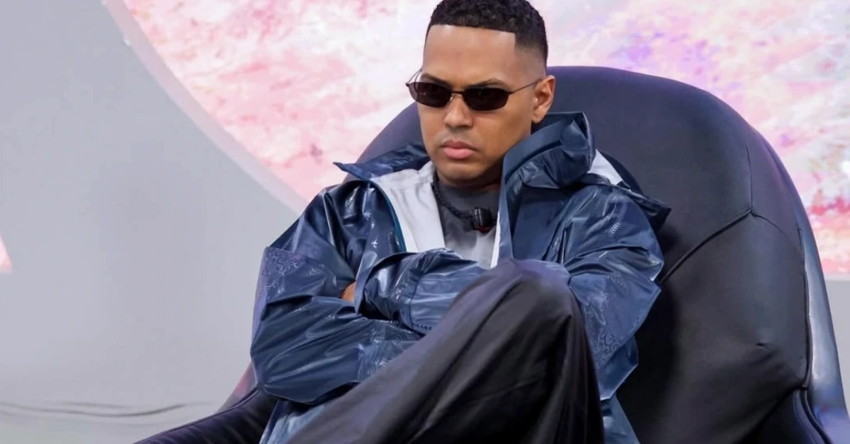 Man wearing dark sunglasses and a blue windbreaker sits with arms crossed in a black lounge chair.