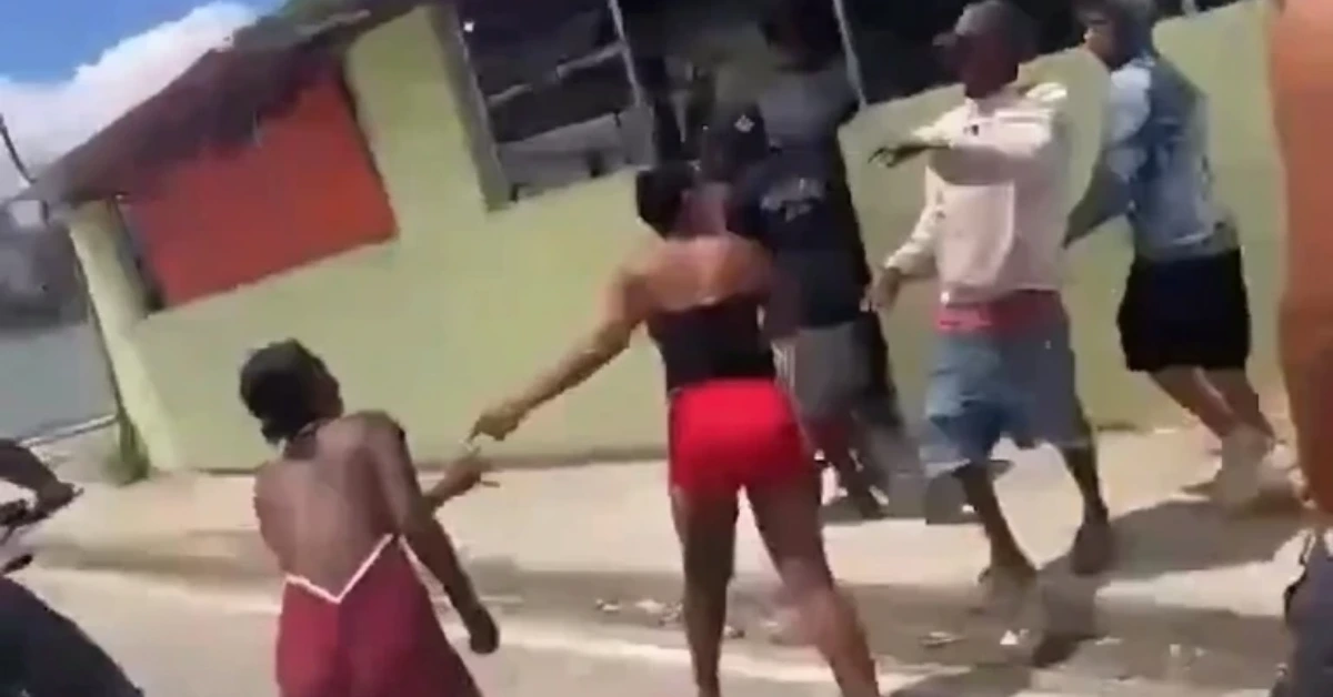 People involved in a street altercation; one person in red shorts reaches toward another in a black top while bystanders watch nearby.