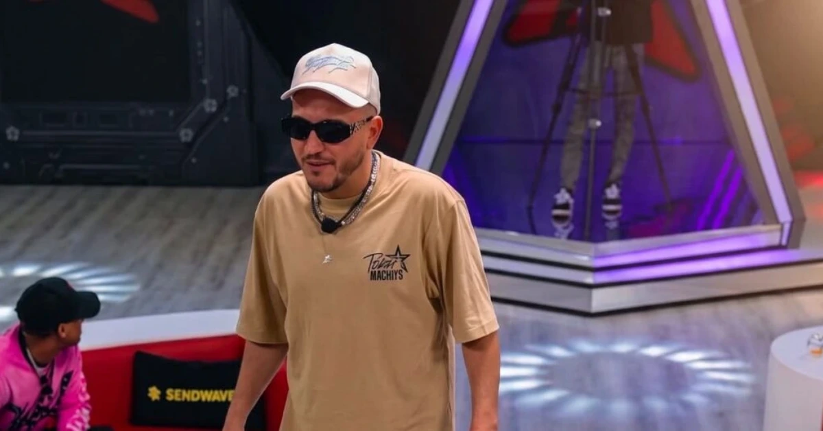 Man wearing a beige shirt, white cap and sunglasses stands in a TV studio with purple lighting and a camera on a tripod in the background.