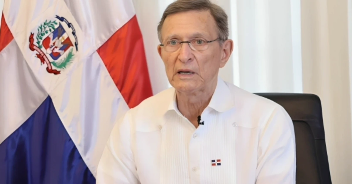 Man with glasses in a white shirt speaking at a desk, Dominican Republic flag in the background.