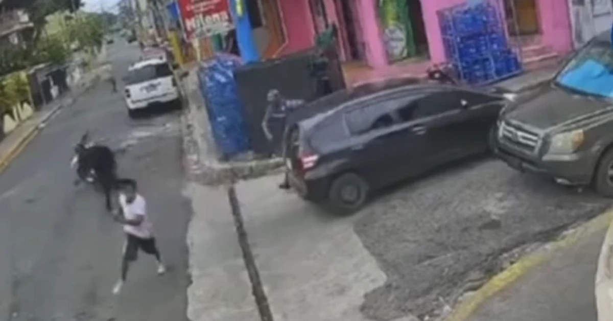Security camera frame of a narrow urban street with colorful storefronts, a dark SUV partially on the curb, and pedestrians nearby. A person in a white shirt runs past on the sidewalk.