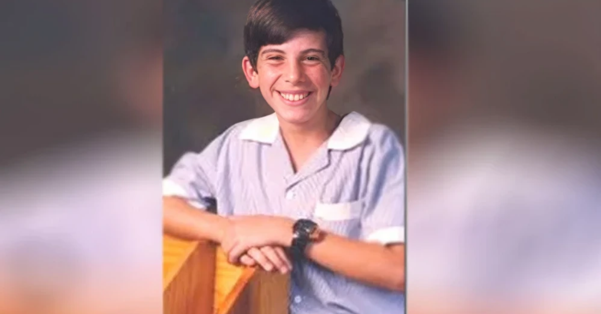 Smiling boy in a light blue striped shirt leaning on a wooden railing, wearing a wristwatch.