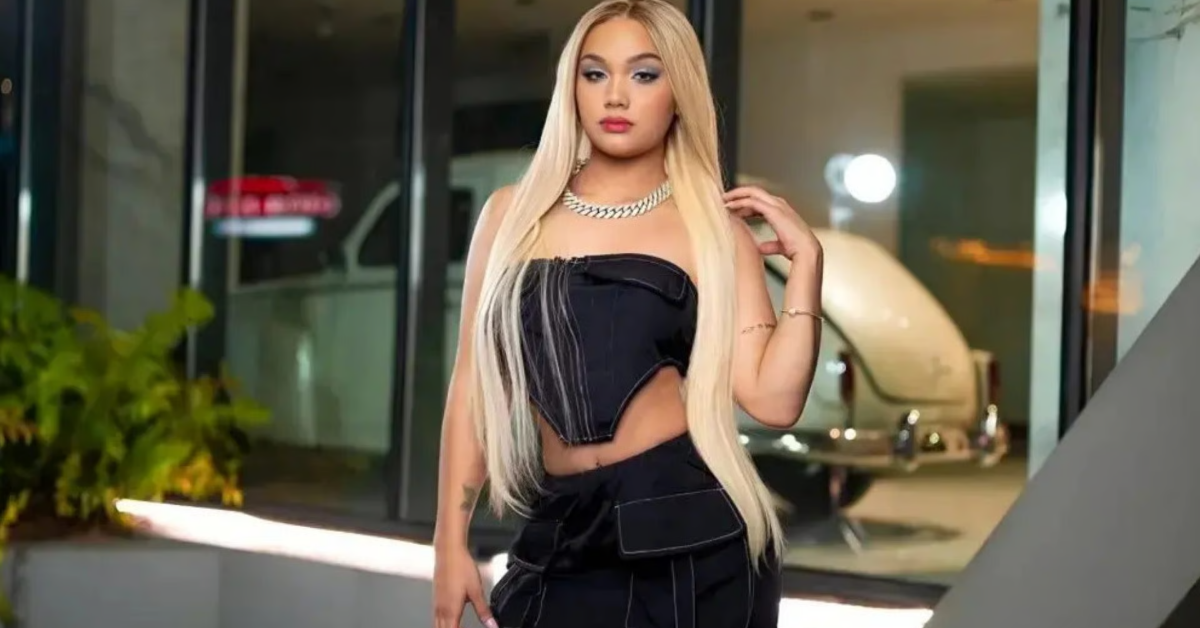 Woman with long blonde hair in a black strapless top and matching pants posing indoors in front of glass doors, wearing a chunky necklace.