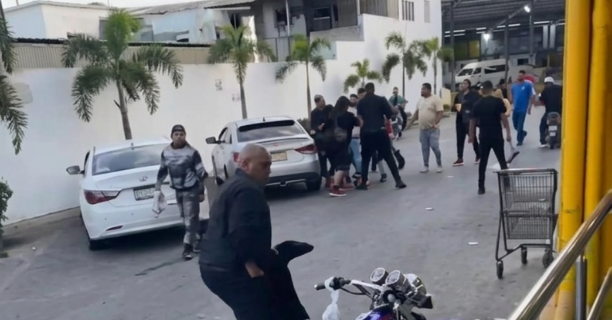Group of people standing and talking in a parking lot beside white cars; a man in the foreground holds a dark bag near a motorcycle.