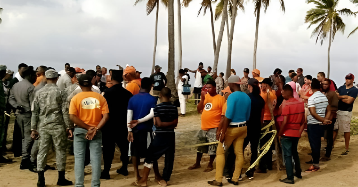 Group of civilians and uniformed personnel forming a circle on a sandy beach, palm trees in the background, around a taped-off area.