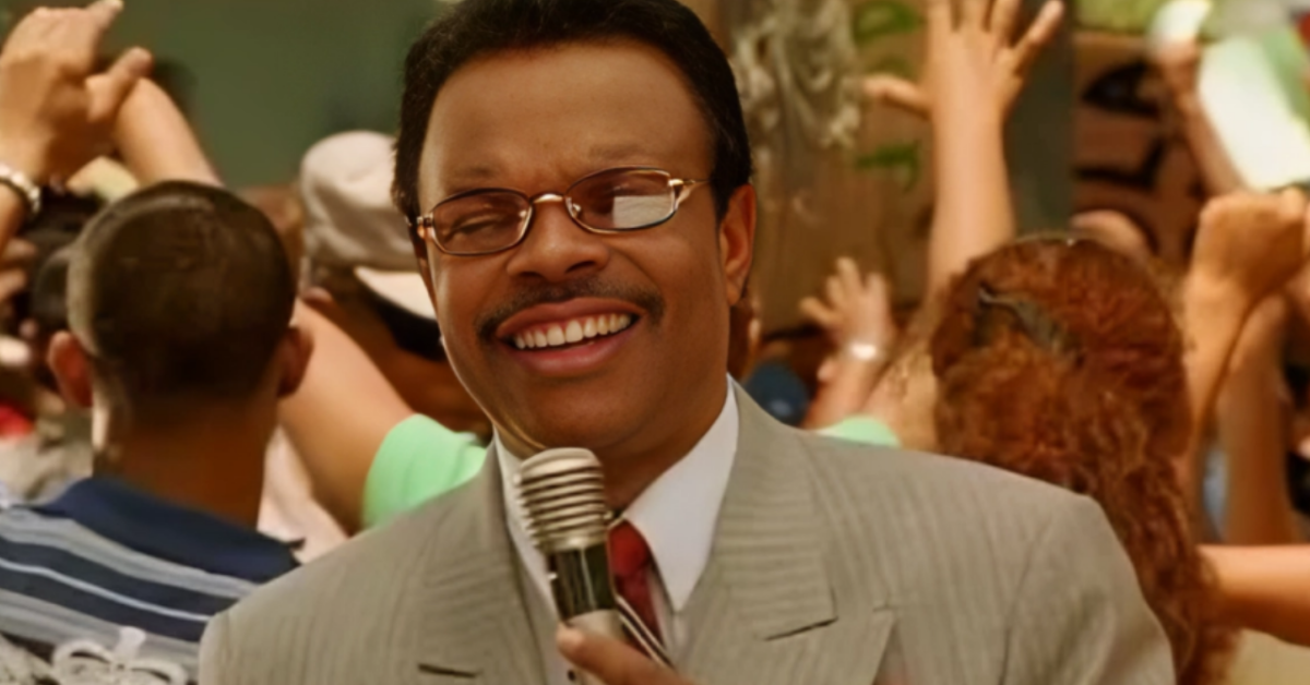 Smiling man in a light gray suit and red tie holding a microphone at a crowded event with raised hands in the background.