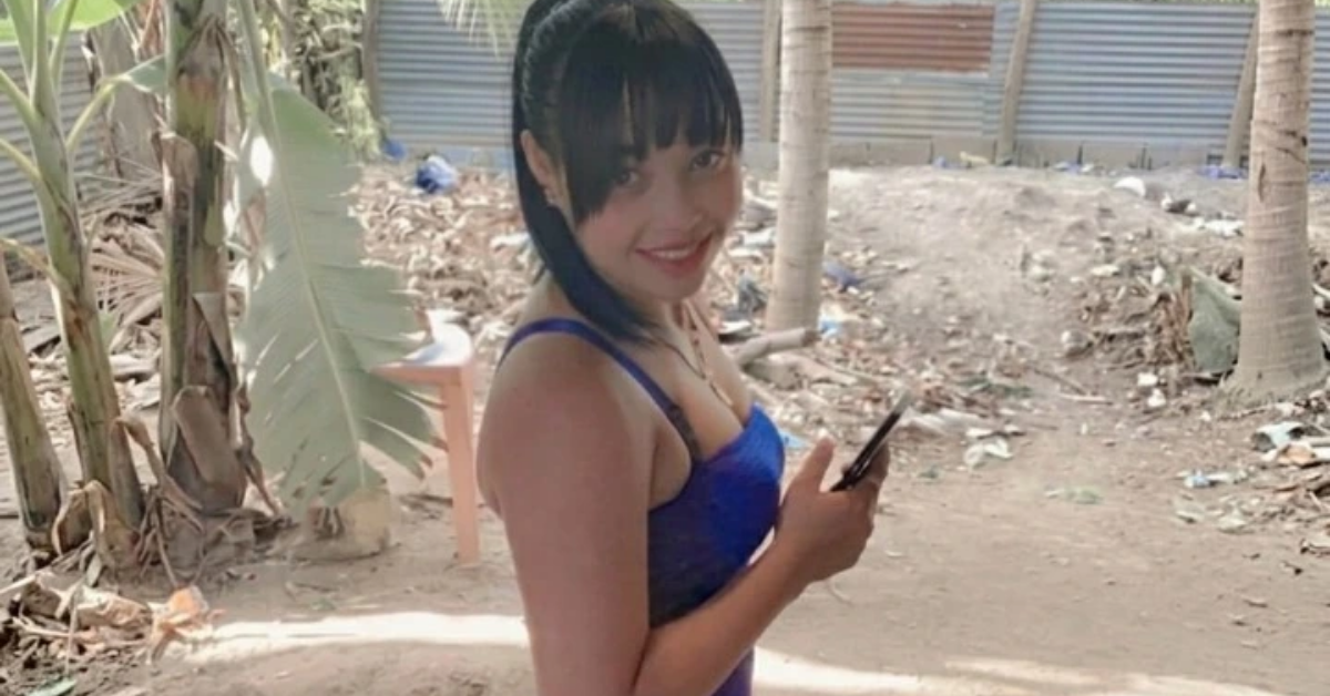 Young woman in a blue top smiles at the camera while holding a smartphone outdoors, with palm trees and a dirt yard in the background.