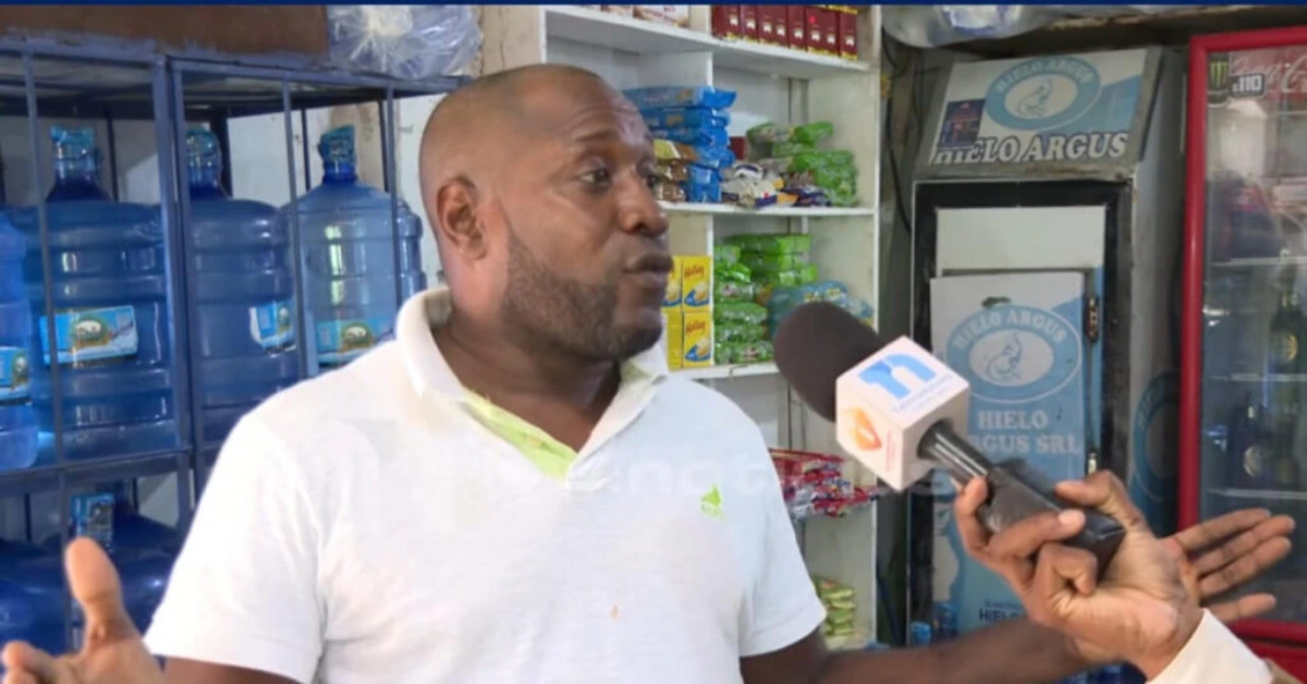 Man in a white polo shirt being interviewed in a shop, with shelves of water jugs and snacks behind him.