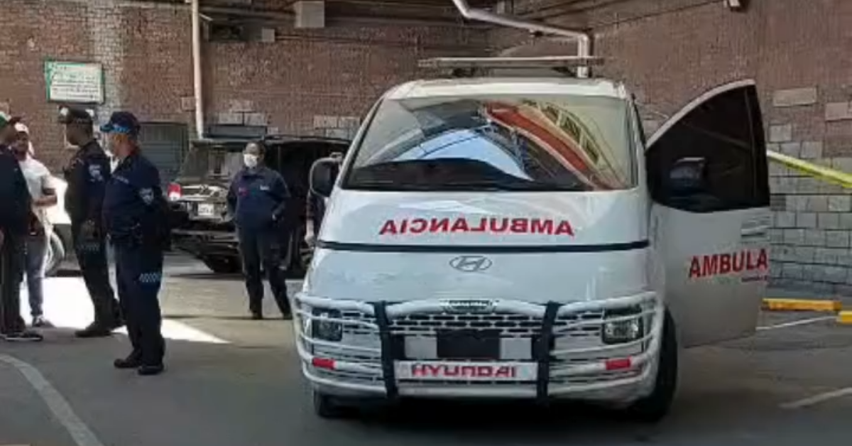 White Hyundai ambulance with front grille guard and open passenger door, 'AMBULANCE' in red on the hood, on a paved lot with uniformed responders nearby.