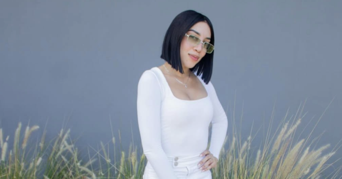 Woman with a black bob haircut and sunglasses poses outdoors in a white outfit, with tall grasses in the background.