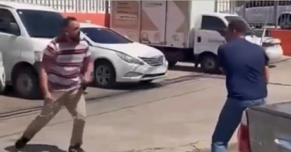 Two men stand in a street facing off, one in a striped polo and khaki pants, the other in a blue T-shirt and jeans, as if about to fight.